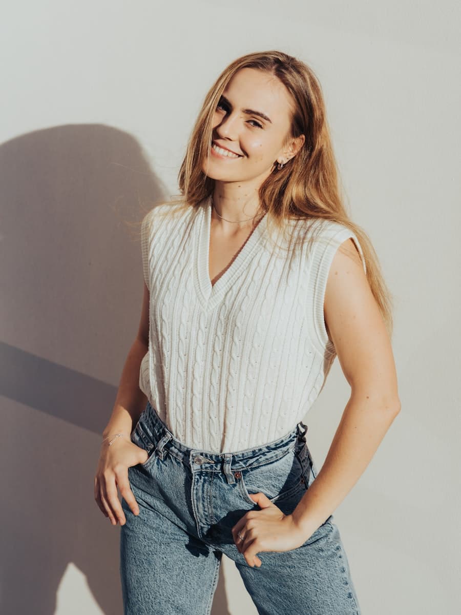 Model wearing a cream cable-knit sleeveless vest with denim