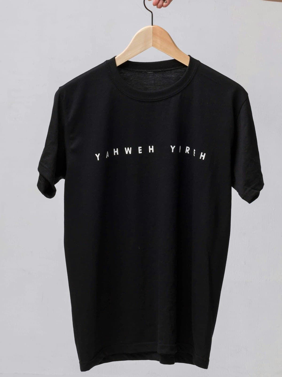 Plain black cotton tee shown on a wooden hanger against a grey wall