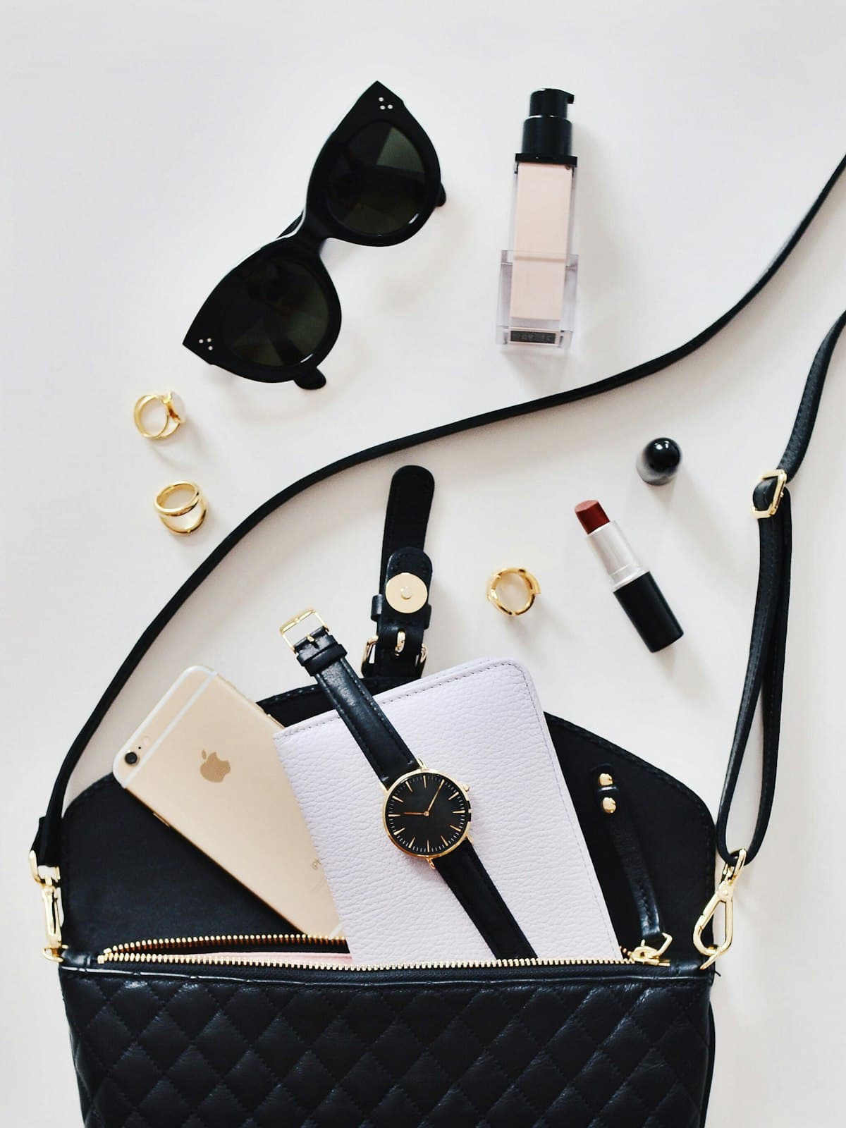 Flat lay of a black quilted crossbody bag with sunglasses, a watch and rings
